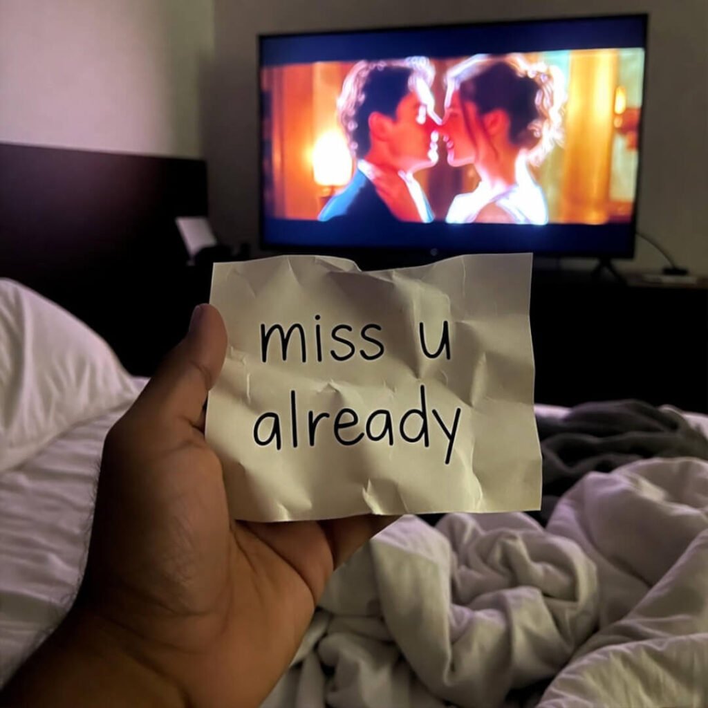 Hand holding "miss u already" note in bed, TV showing kissing couple.