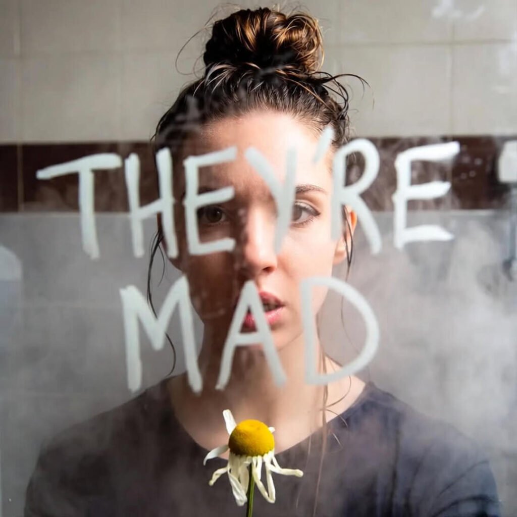 Foggy mirror selfie: messy bun, wilted daisy, "THEY'RE MAD" scrawled in marker.